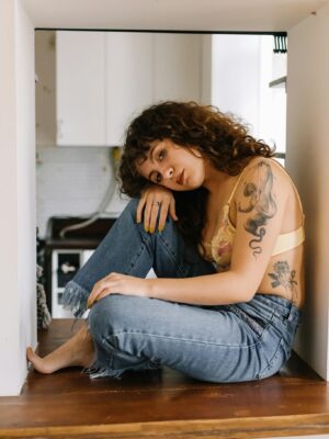 Curly-haired woman with tattoos sits in a cozy indoor space wearing denim.